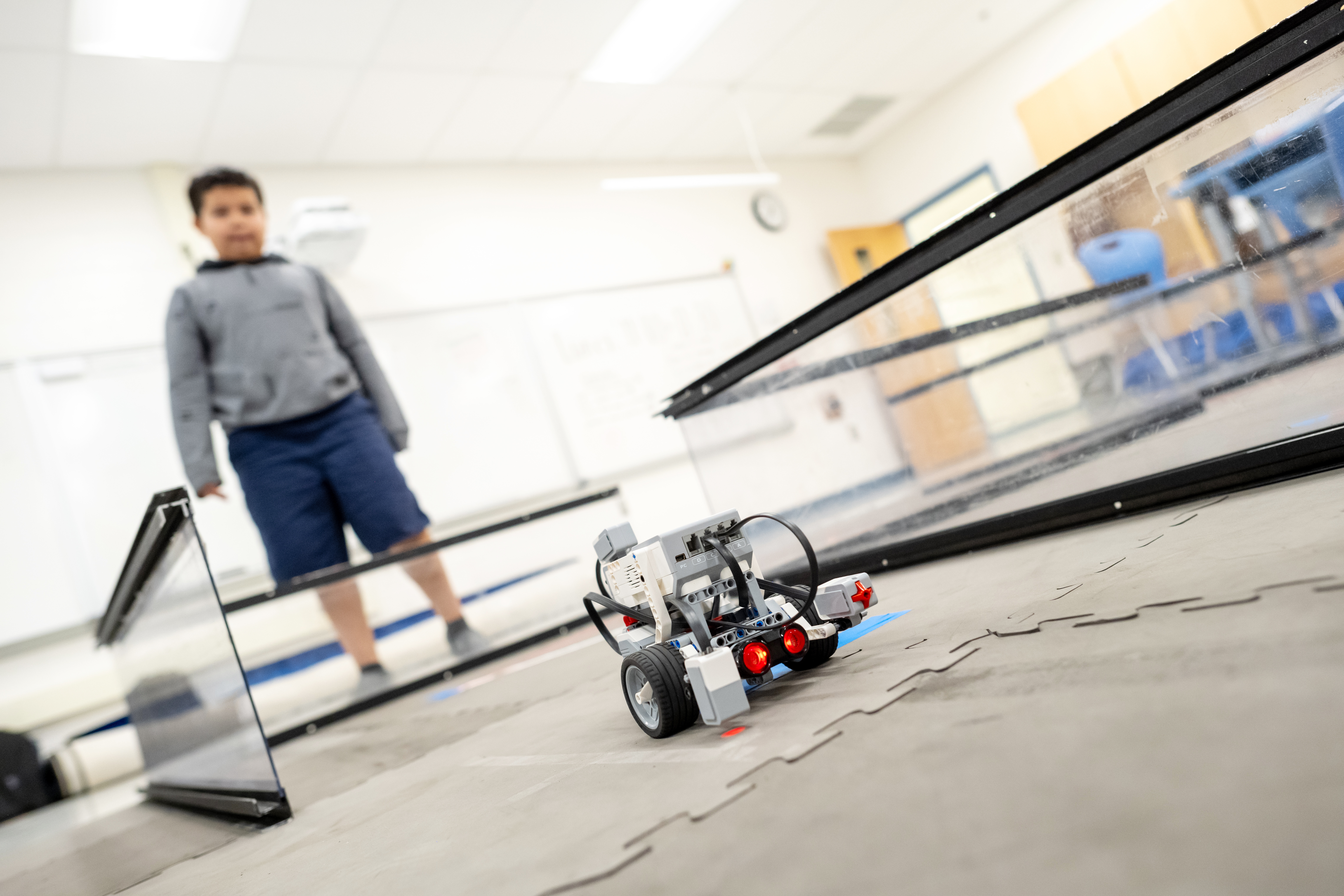 A student watches as a LEGO robots rolls through glass wall corridors on the floor. Red lights shine at the front of the robot.