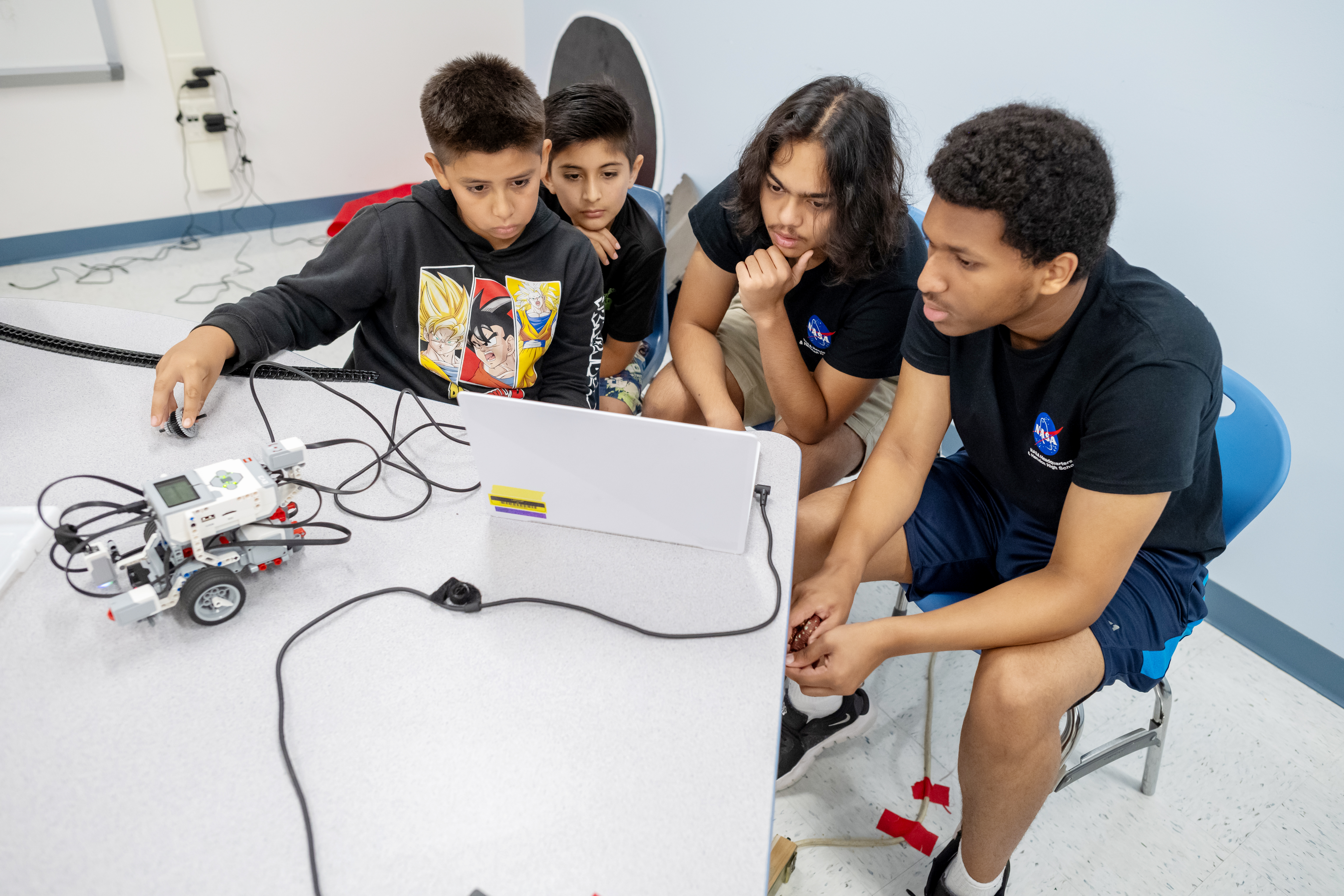 Two students from Herndon High School, right, and two Clearview students, left, look at a laptop which the students are using to program a robot. The robot, resting left of the computer, is connected to the computer by a cable. One Herndon student scratches his chin as he looks closely at the computer screen. 