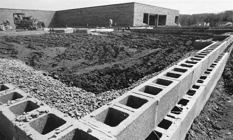 Black and white photograph, taken in 1982, showing the new wing under construction. Work has just begun on the cinderblock walls and the grounds inside are being graded in preparation for the pouring of the concrete pad. The existing building and a bulldozer can be seen in the background.