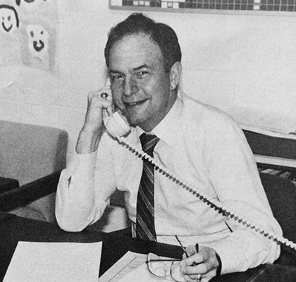 Black and white photograph of Bernie Gross from our 1987 yearbook. He is seated at his desk, talking on the phone. 